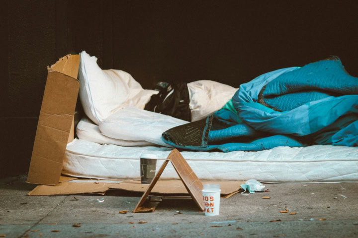 Persoon ligt op een matras op straat. Ernaast staat een bordje en bekertje om te bedelen.