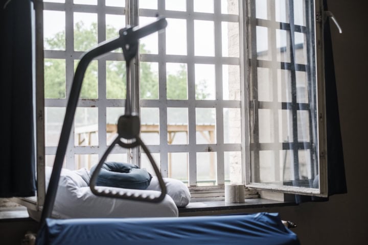 Een bed op de ouderenafdeling in de gevangenis van Merksplas met daarachter een raam met tralies.